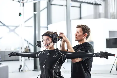 Schauspielerin im Motion-Capture-Anzug mit Kopfkamera, ein Techniker justiert die Ausrüstung